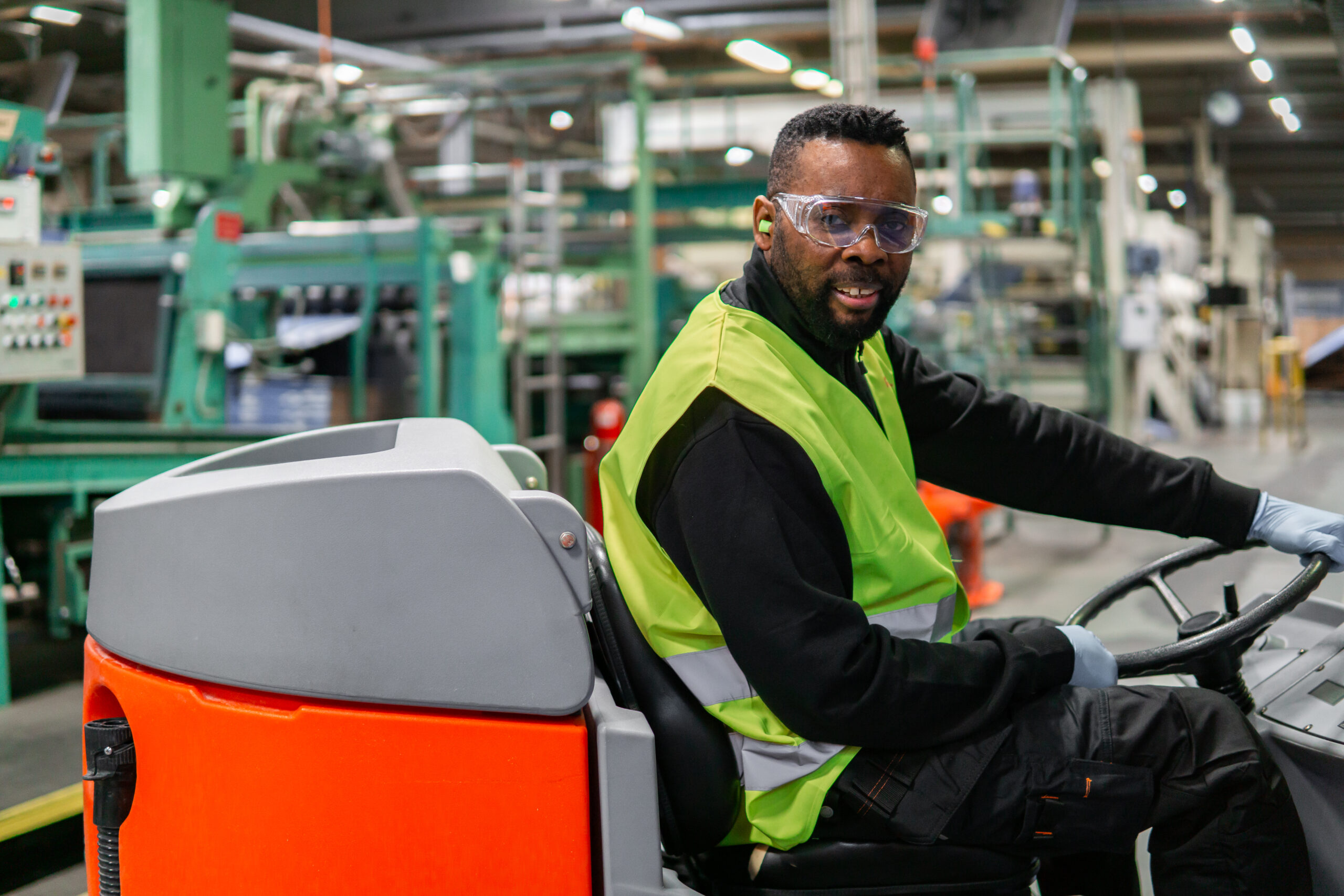 Arbetare i säkerhetsväst och skyddsglasögon kör städmaskin i en industrimiljö.